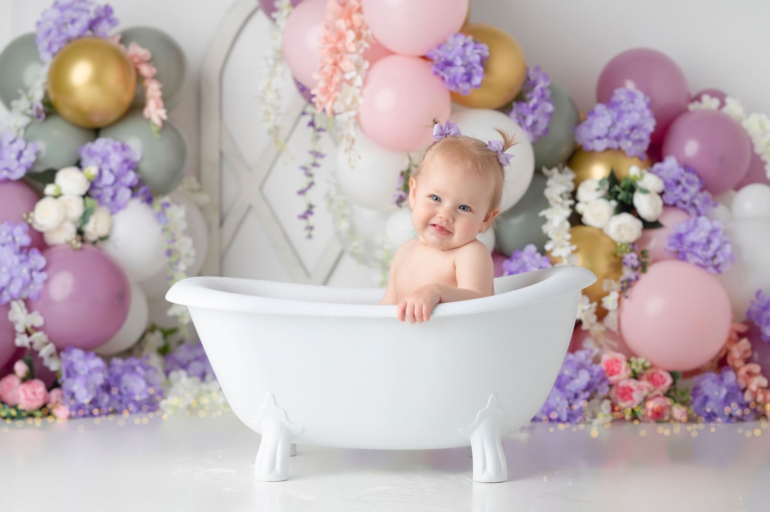 Baby girl sitting in a white clawfoot bathtub surrounded by colorful balloons and flowers for a first birthday cake smash.