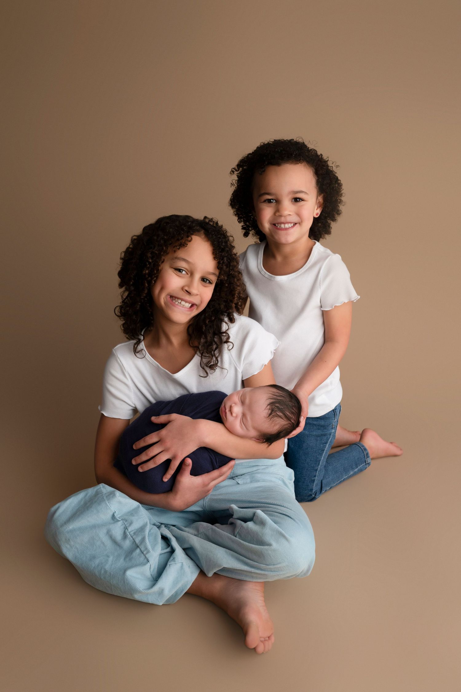 Two smiling girls in white shirts hold a sleeping newborn baby wrapped in navy blue against a tan background.