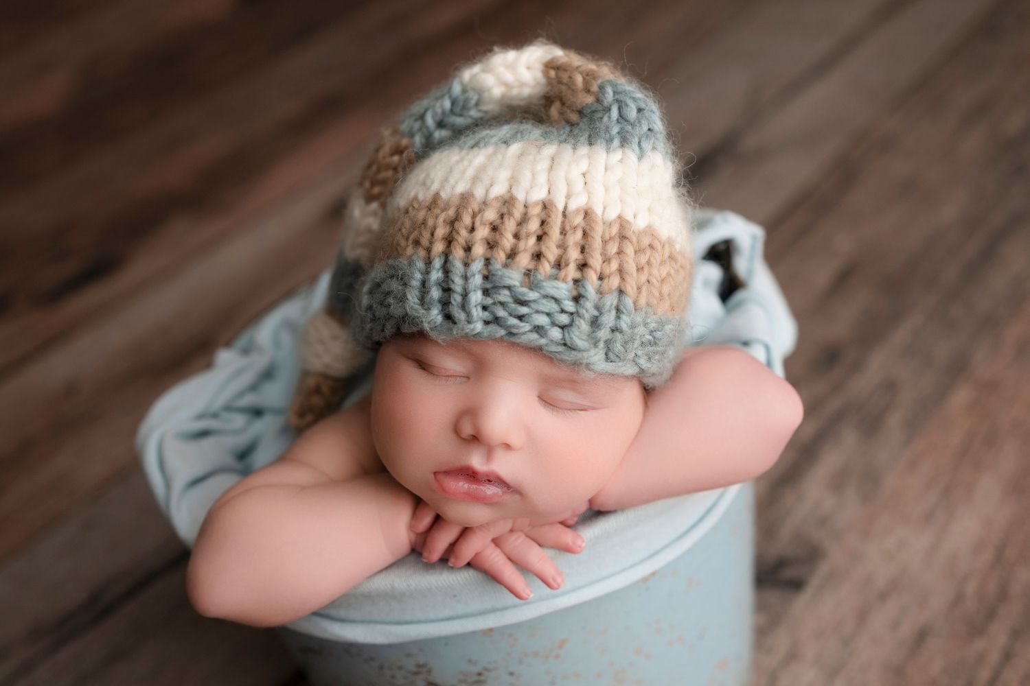 Sleeping newborn baby wearing a striped knit hat resting in a blue bucket on a wooden floor.