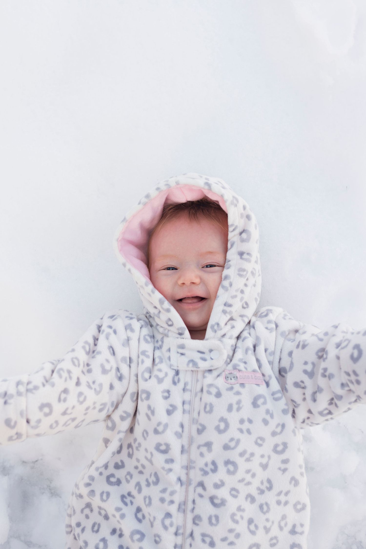 A smiling baby in a white leopard print snowsuit with pink hood lining lying in snow.