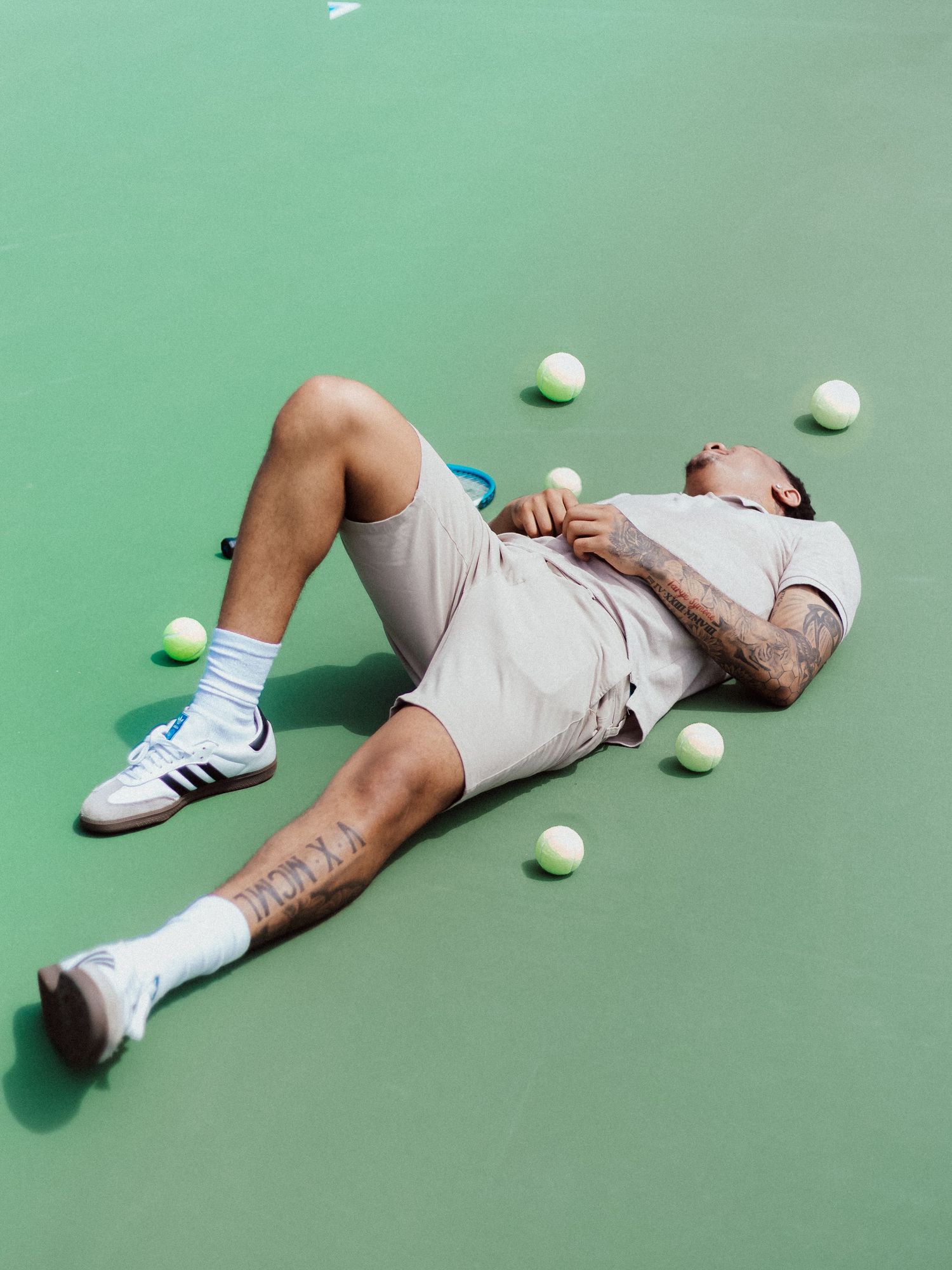 Tennis player lying exhausted on green court surrounded by tennis balls wearing white outfit and sneakers.