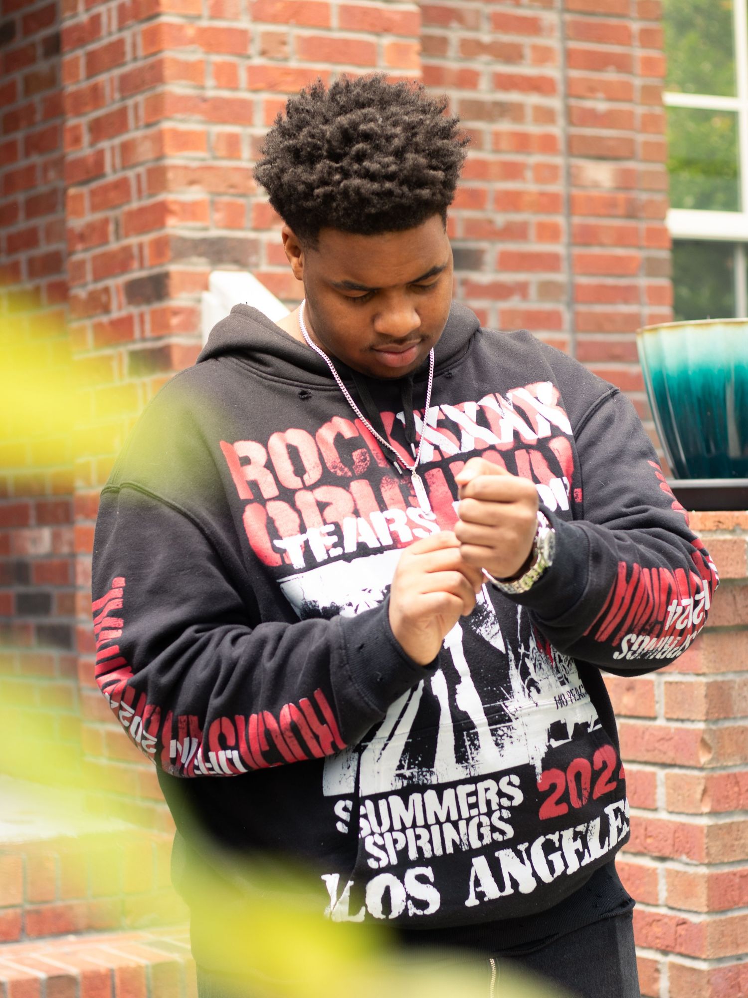 A person in a black hoodie with graphic text stands against a brick wall background.