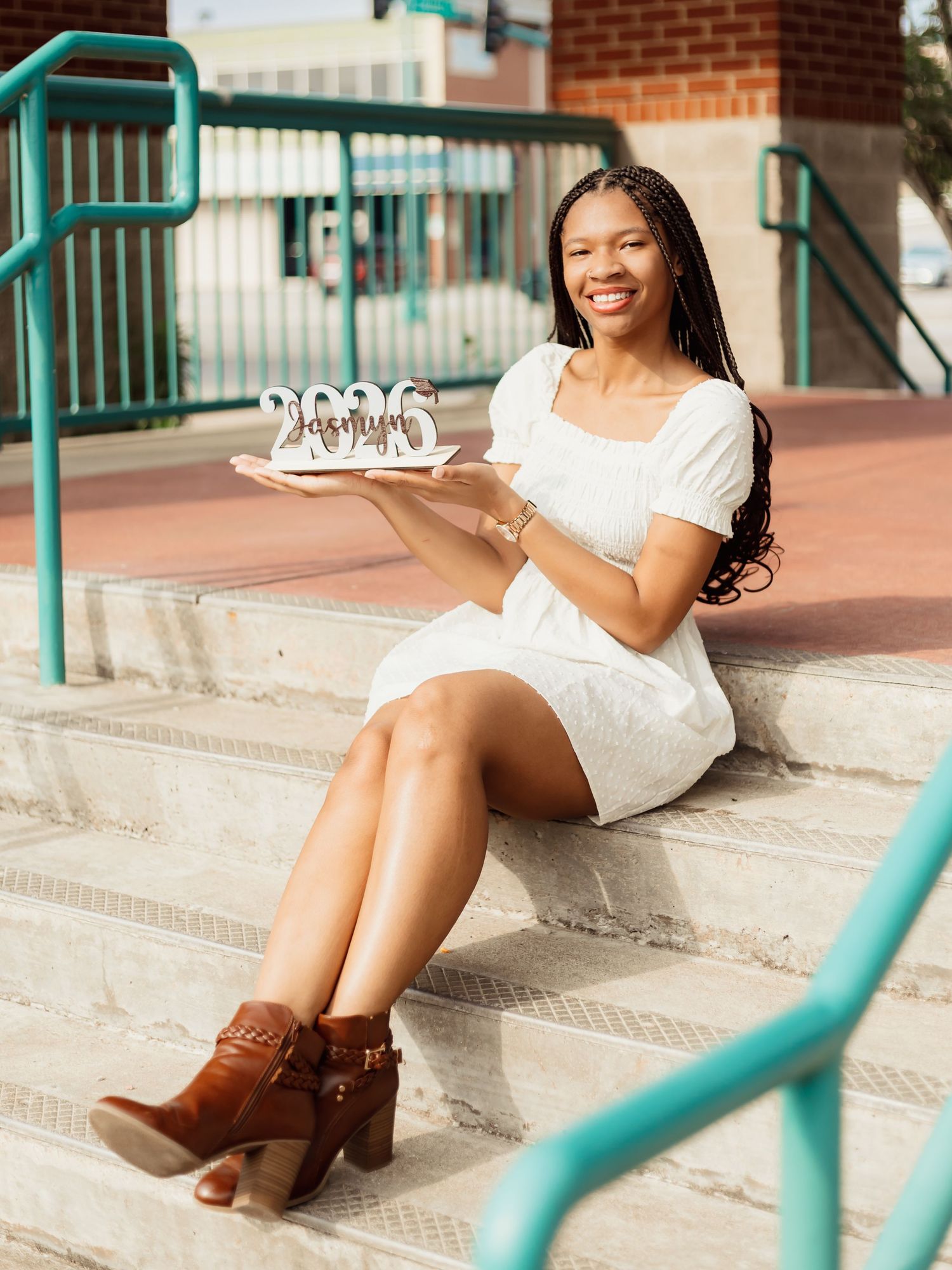 A person in a white dress sits on outdoor steps holding a birthday cake with candles.