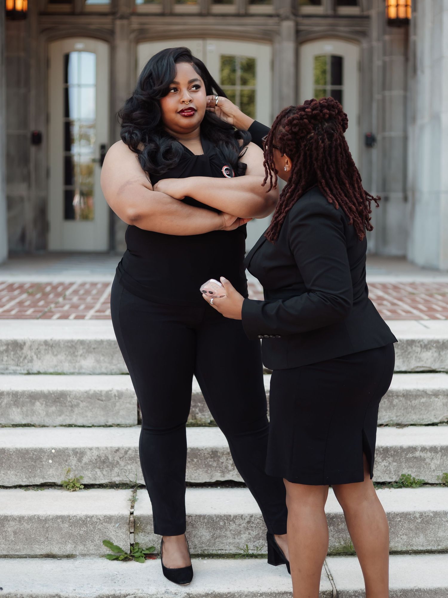 Two people in black outfits share playful poses on concrete steps outside a building with arched architecture.