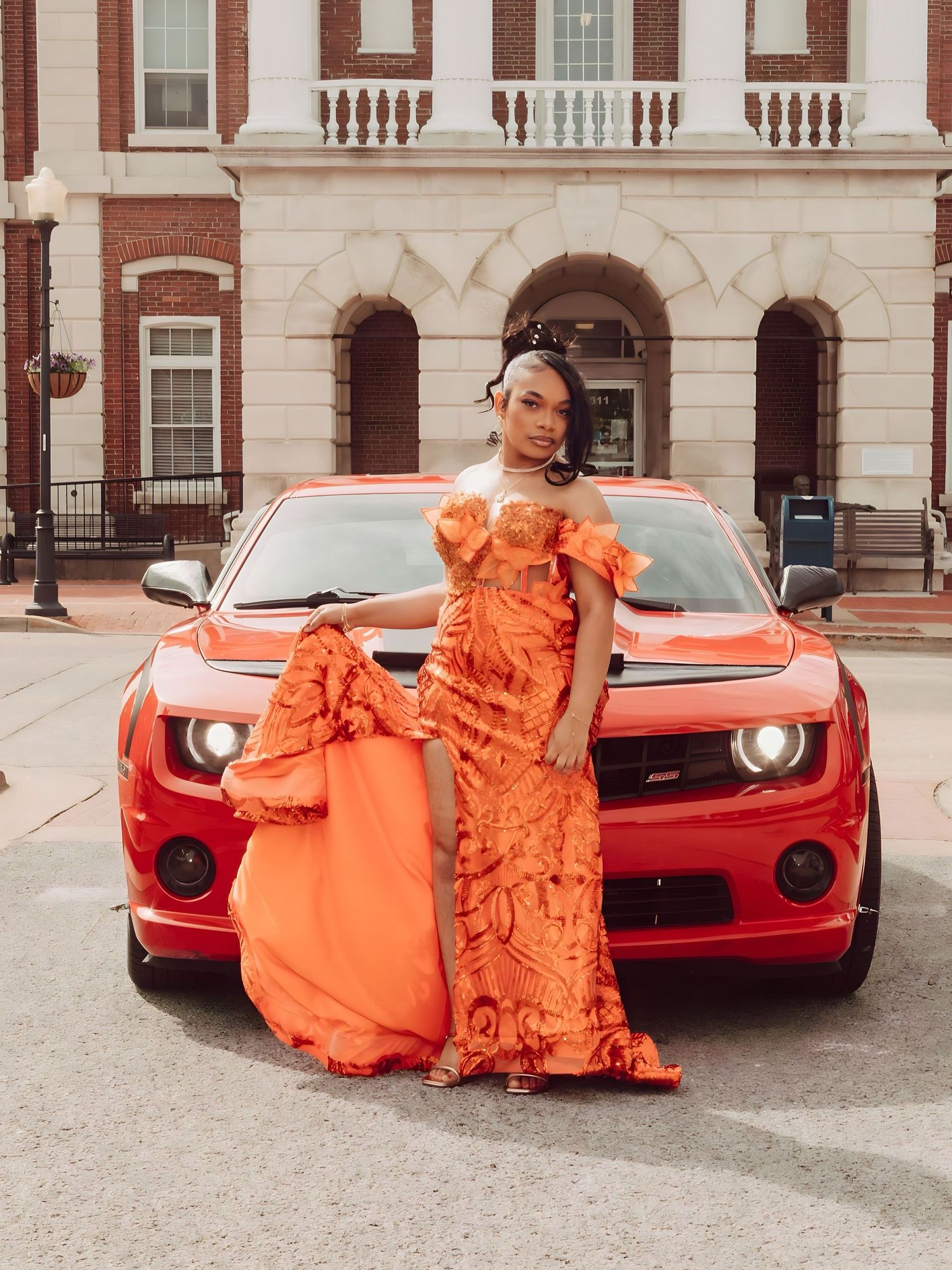 A vibrant orange ruched gown is showcased against a red sports car in front of a classic brick building.