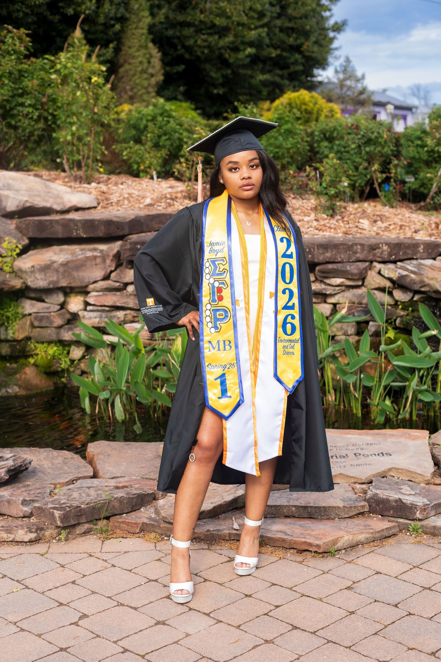 Young woman in black graduation gown and 2026 stole poses proudly outdoors near a stone wall and pond.