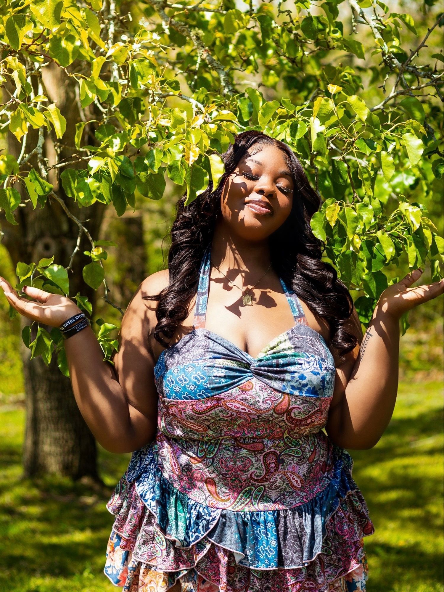 A woman in a patterned sundress stands joyfully with raised arms among lush green trees on a sunny day.