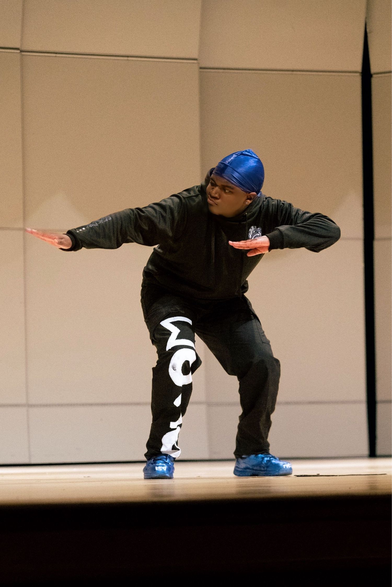 Performer in black outfit with blue hat and shoes holds red props while dancing on stage.