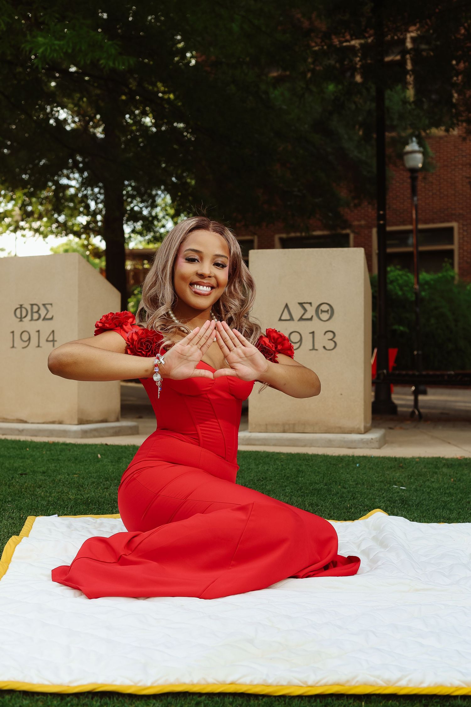 Woman in red dress sitting on white blanket making hand sign in front of Greek fraternity monument stones.