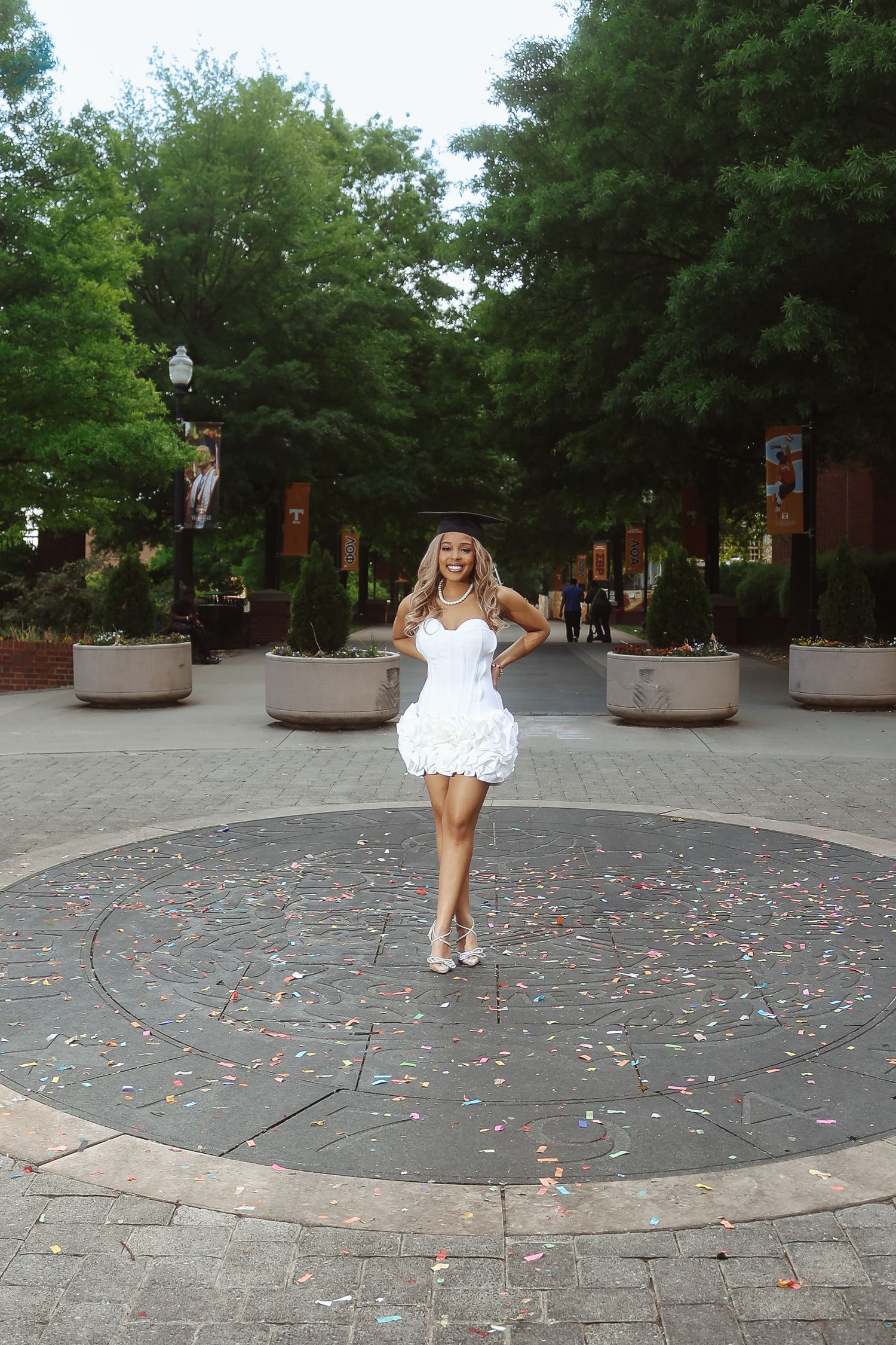 Woman in white mini dress posing confidently on a decorative circular plaza surrounded by lush green trees.