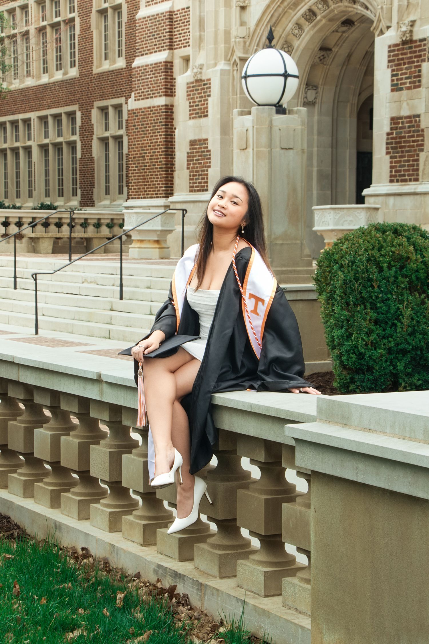 A smiling young woman in graduation cap and gown sits on a stone railing outside a brick university building.