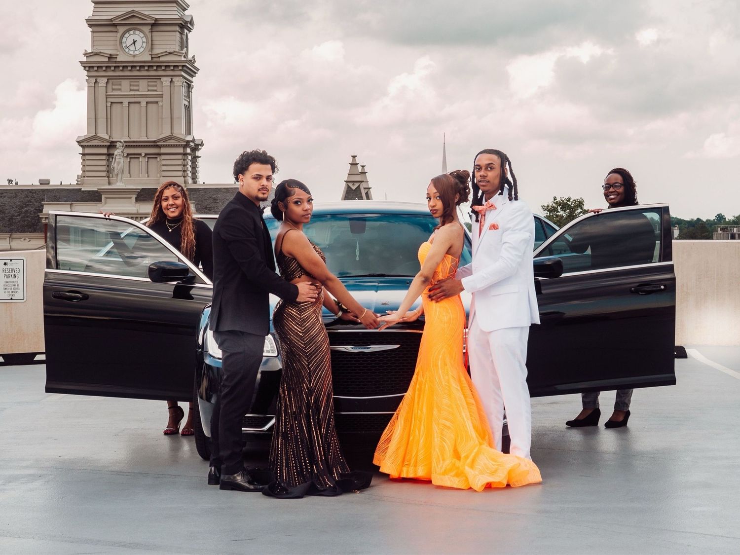 People in formal attire gather around luxury vehicles for a special occasion photo.