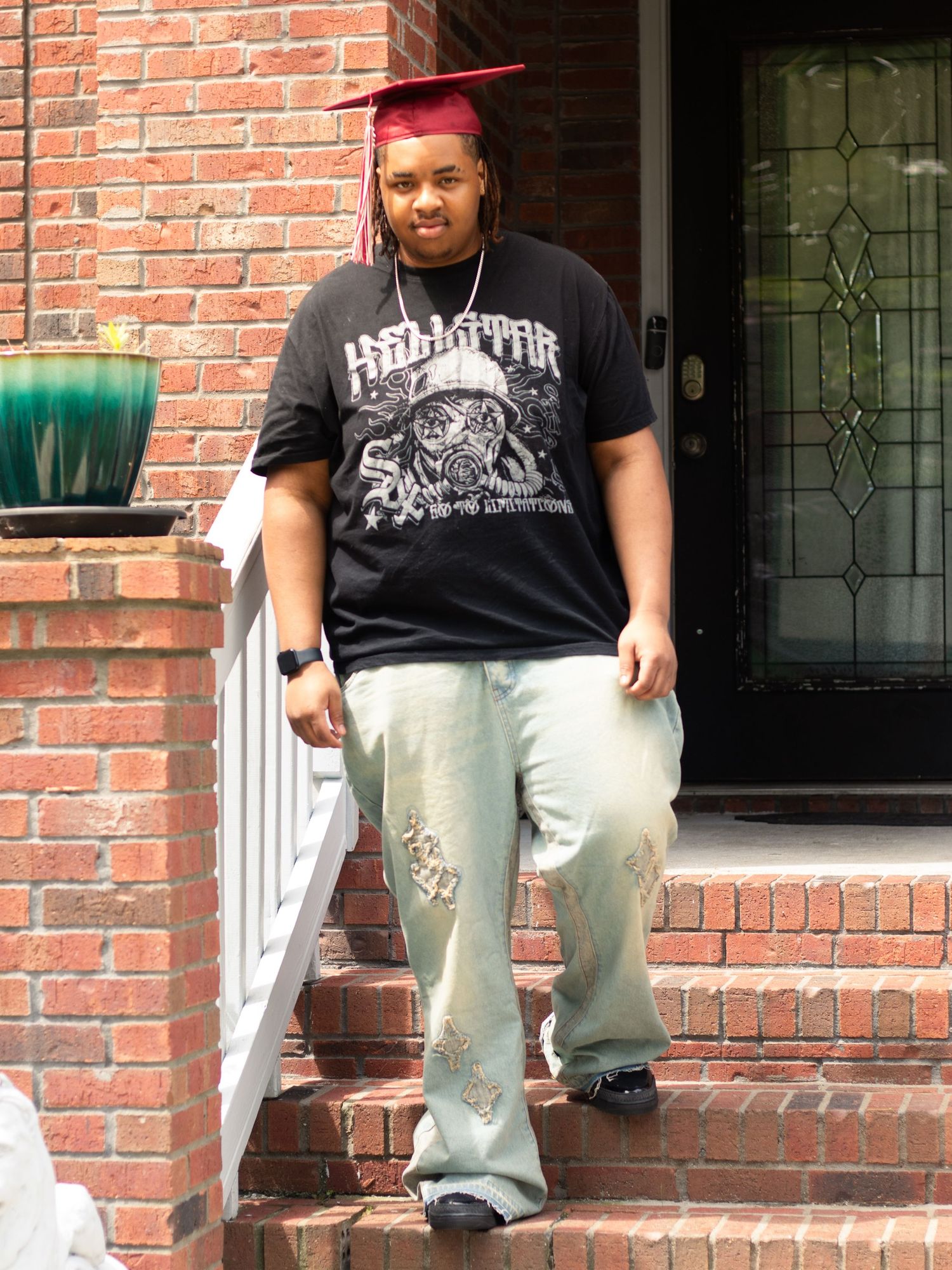 A graduate in a black t-shirt and light pants poses on brick steps wearing a red graduation cap.