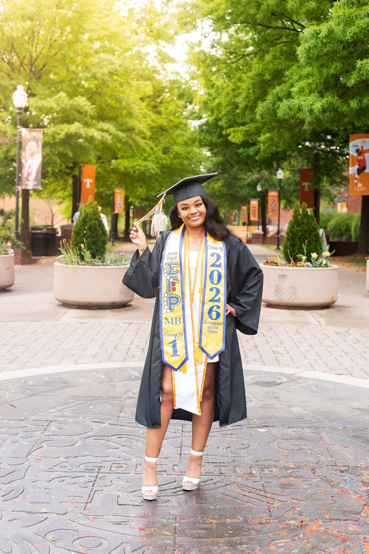 Smiling graduate in black cap and gown with blue and gold 2026 stole poses outdoors on a sunny campus plaza.