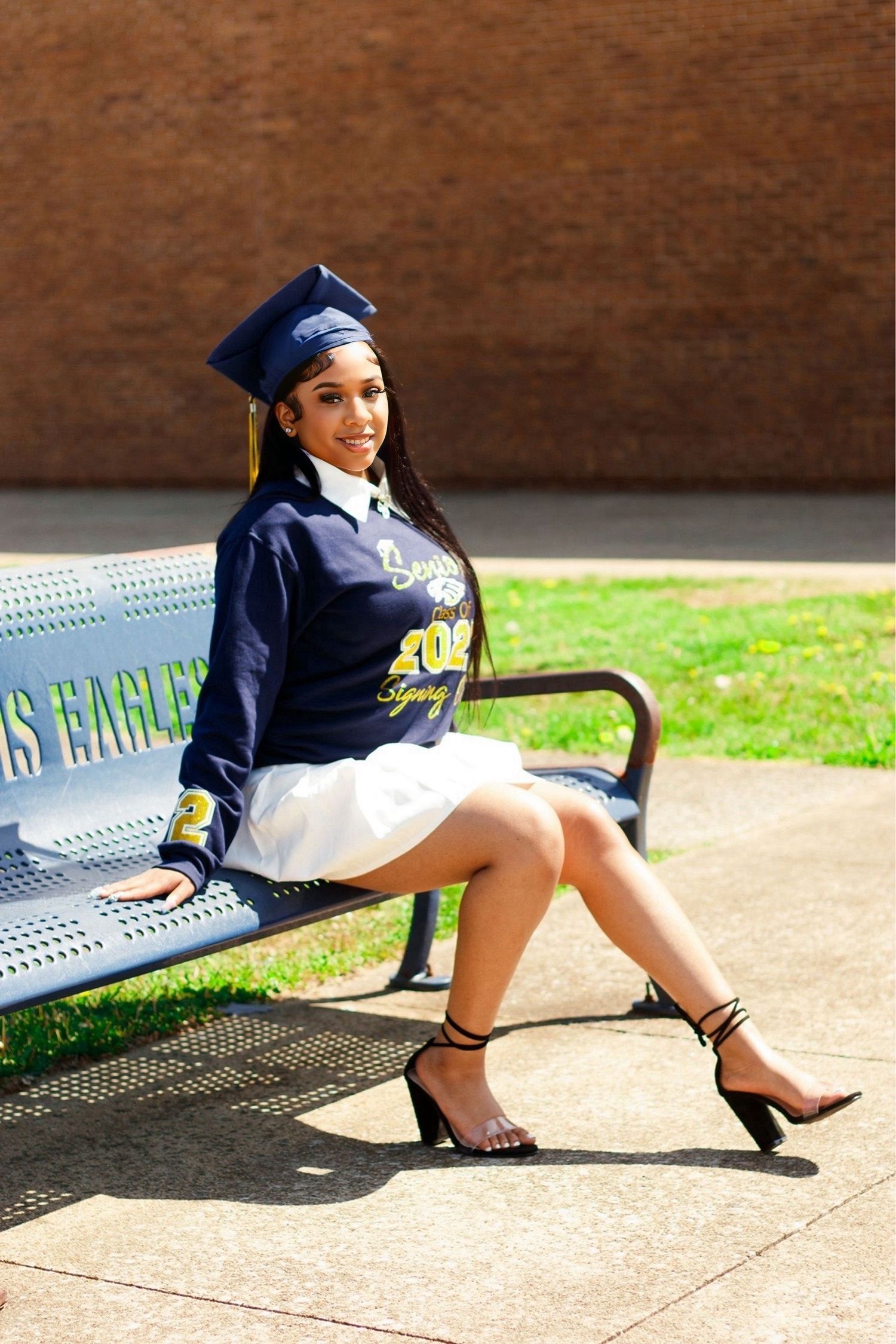 Graduate in navy cap and gown poses on metal bench during outdoor photoshoot.