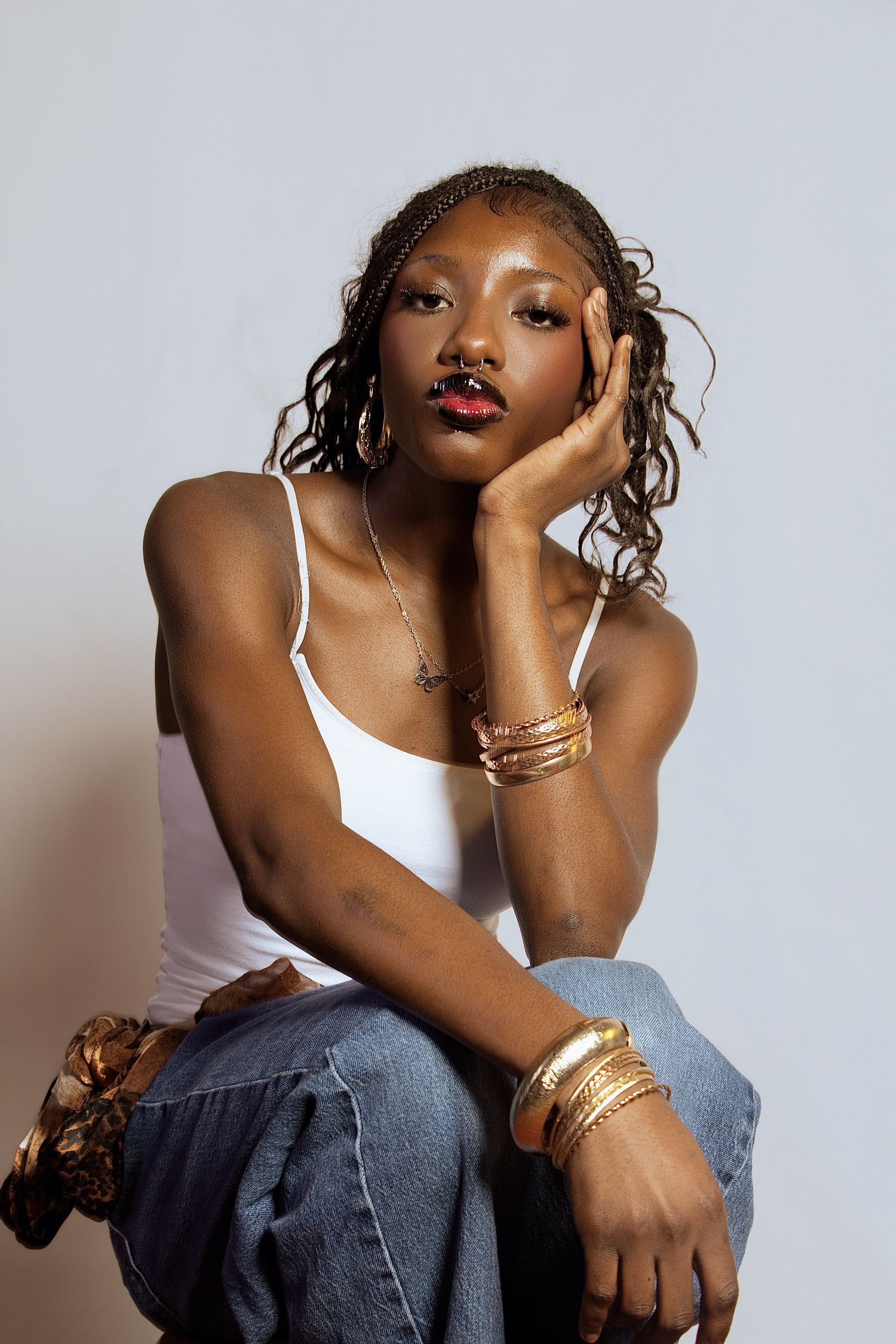 Fashion portrait of model in white tank top and jeans wearing gold bangles against neutral background.