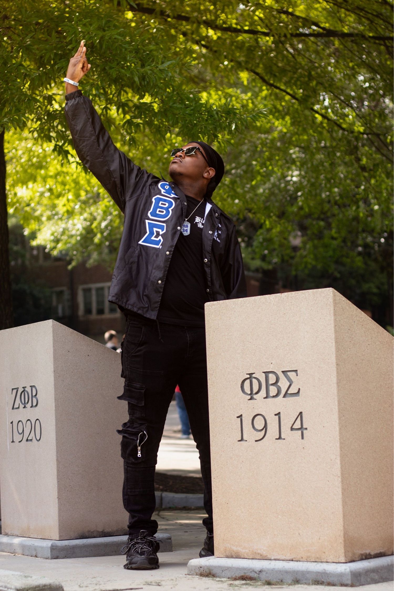 Man in Phi Beta Sigma jacket poses between ΖΦΒ 1920 and ΦΒΣ 1914 fraternity monuments outdoors.