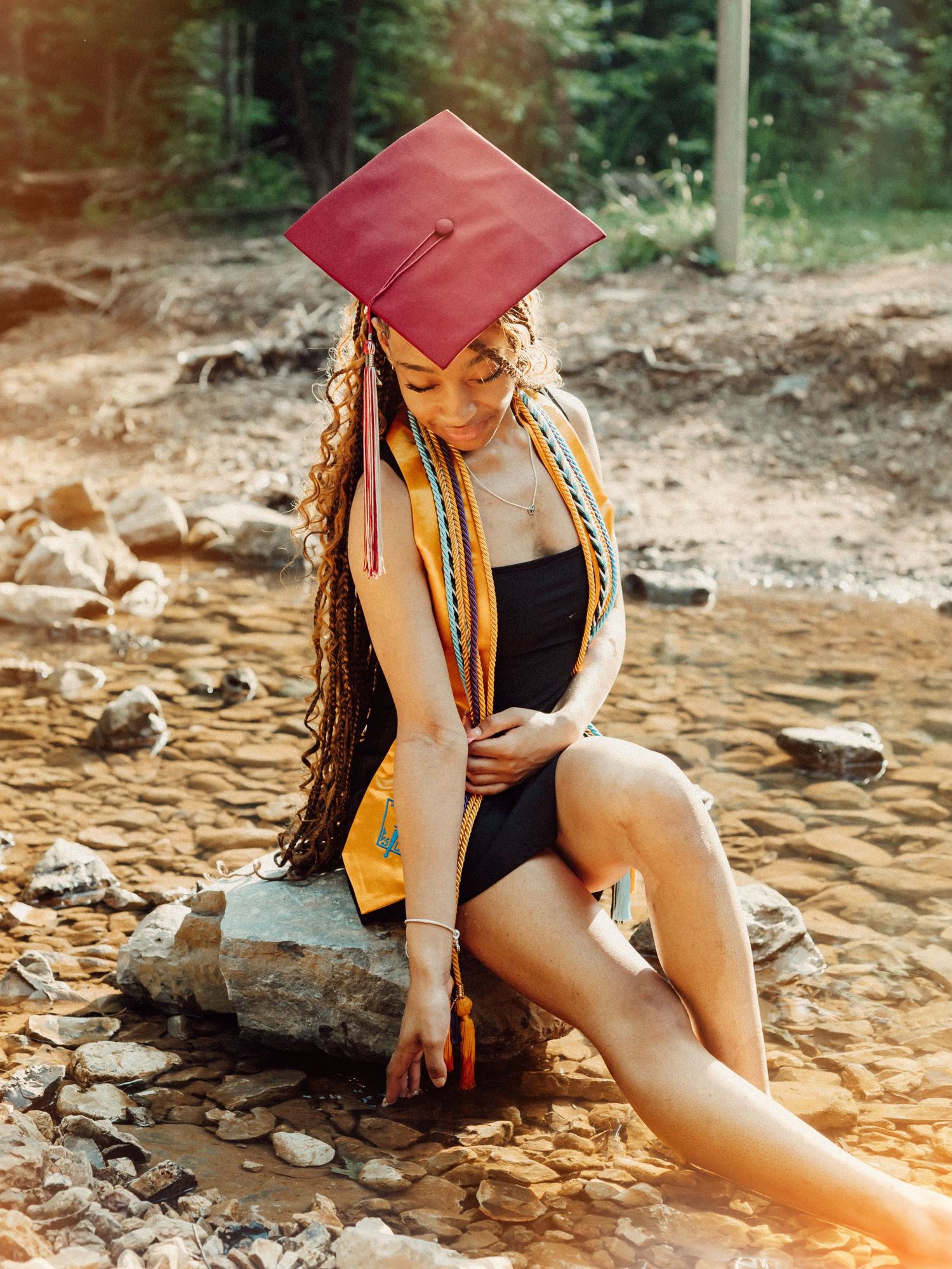 Graduate in red cap and honors cords sits on rocky outcrop in nature during outdoor graduation photoshoot.
