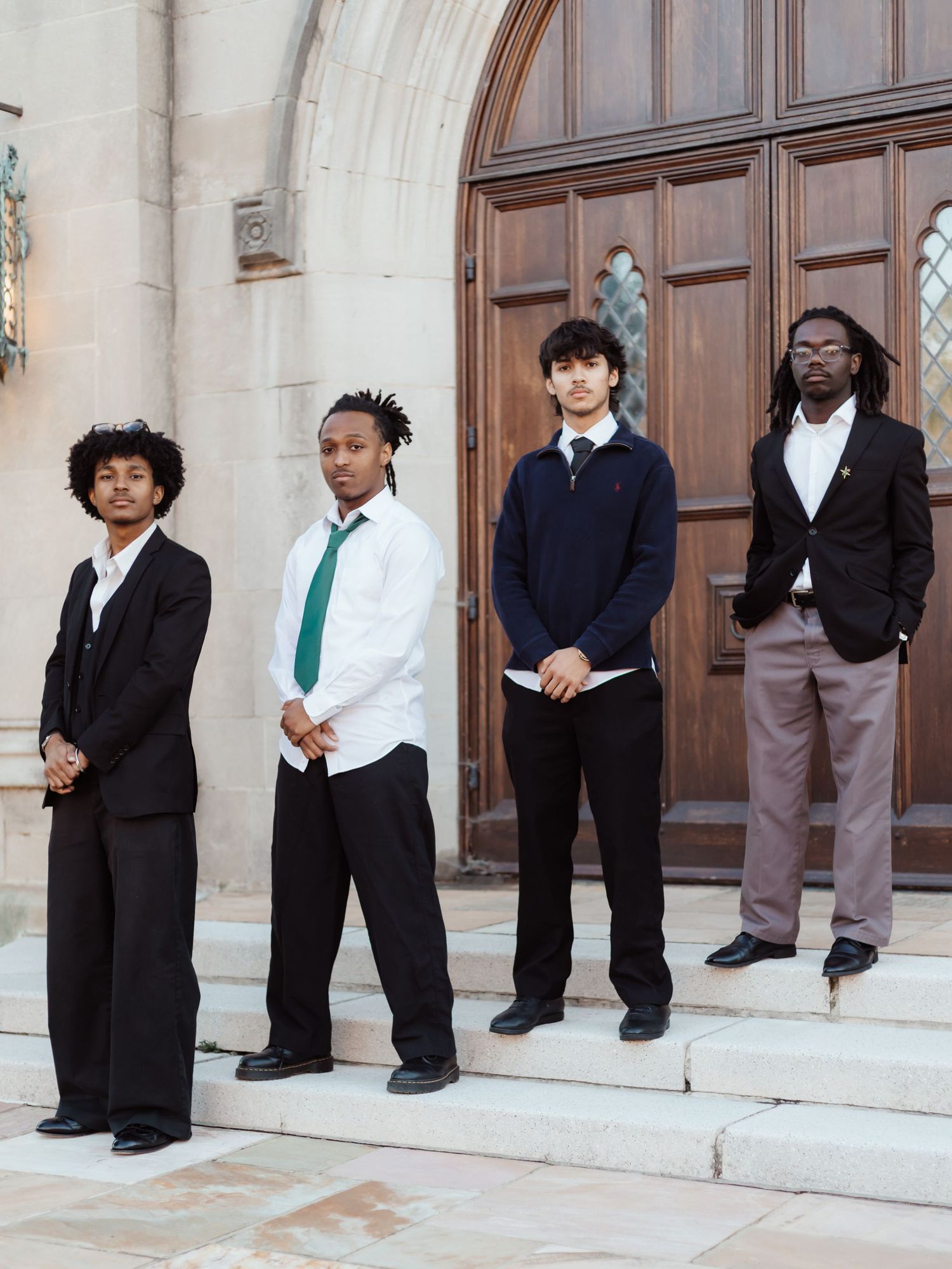 A wedding party poses on church steps with groomsmen in dark suits and bridesmaids in teal dresses.