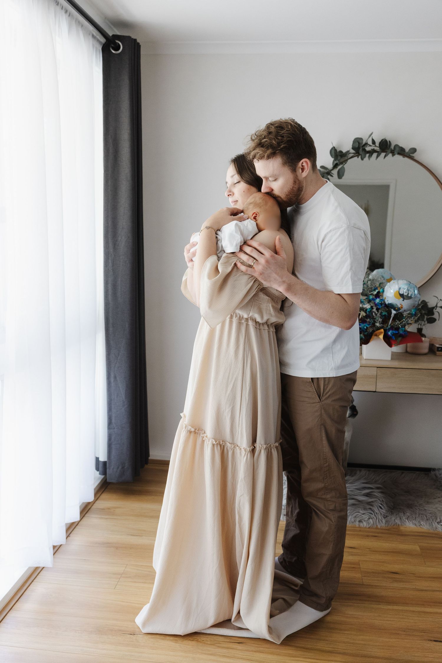 A new mother and father cradle their newborn baby near a window in a Canberra newborn photoshoot