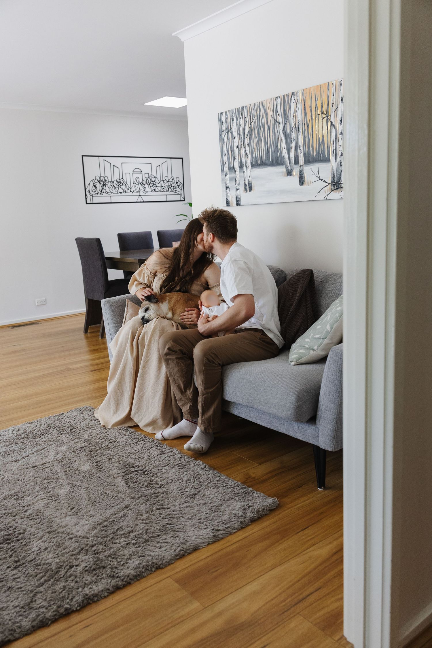 A couple kisses on the couch with their baby and dog in their newborn photoshoot in Canberra