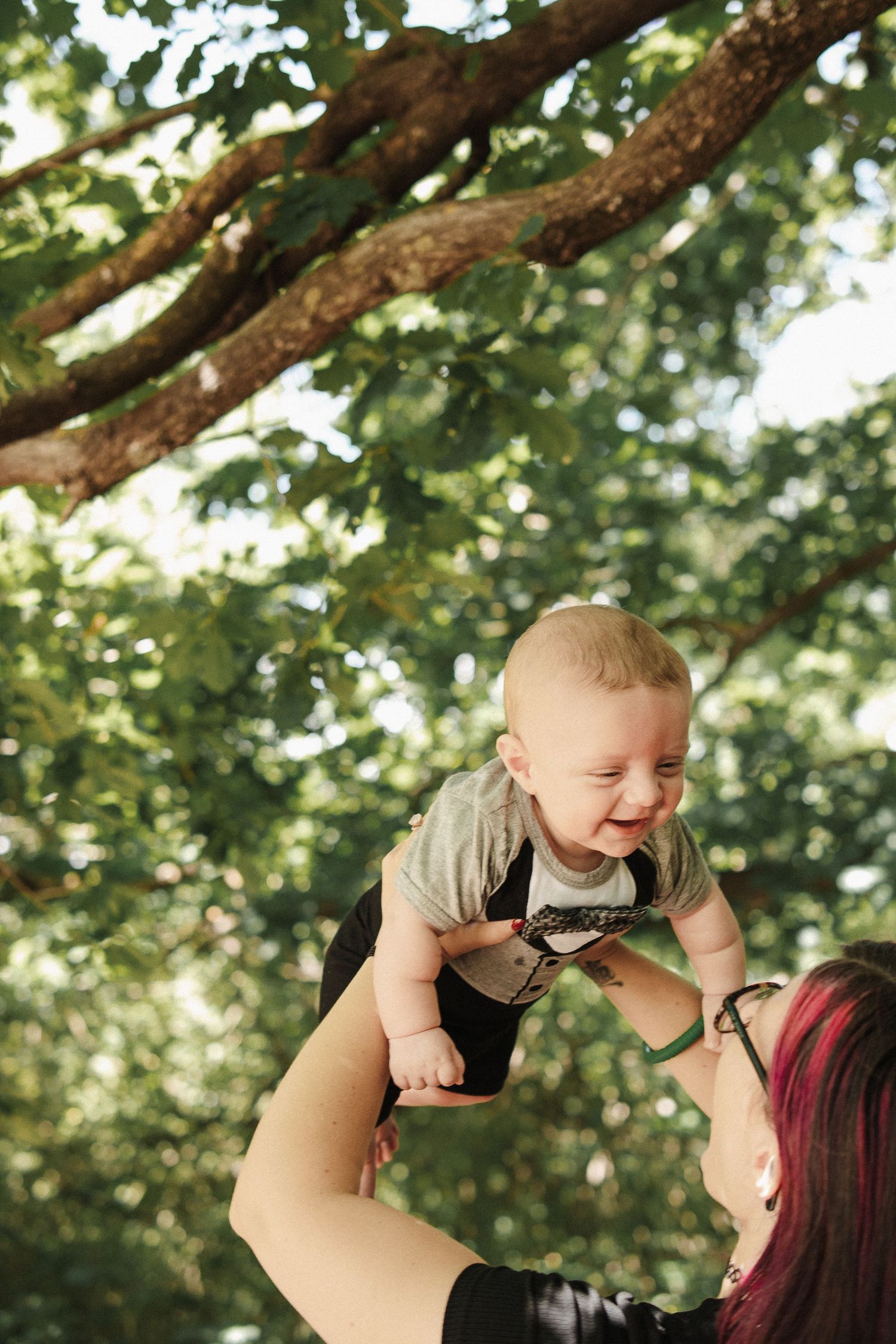 A mother holds her laughing baby above her head against a backdrop of green trees, in a Canberra family photoshoot