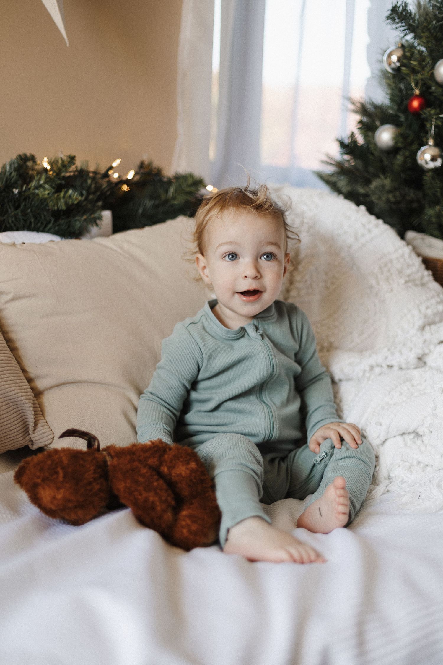 A toddler sits on a white couch with a teddy and a christmas tree in a Canberra child studio photoshoot