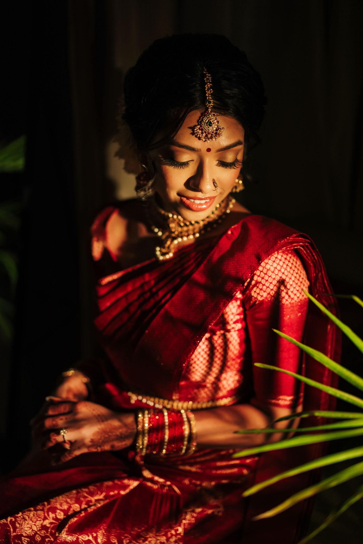 A bride in a red silk saree with gold jewelry and maang tikka poses gracefully in warm light.