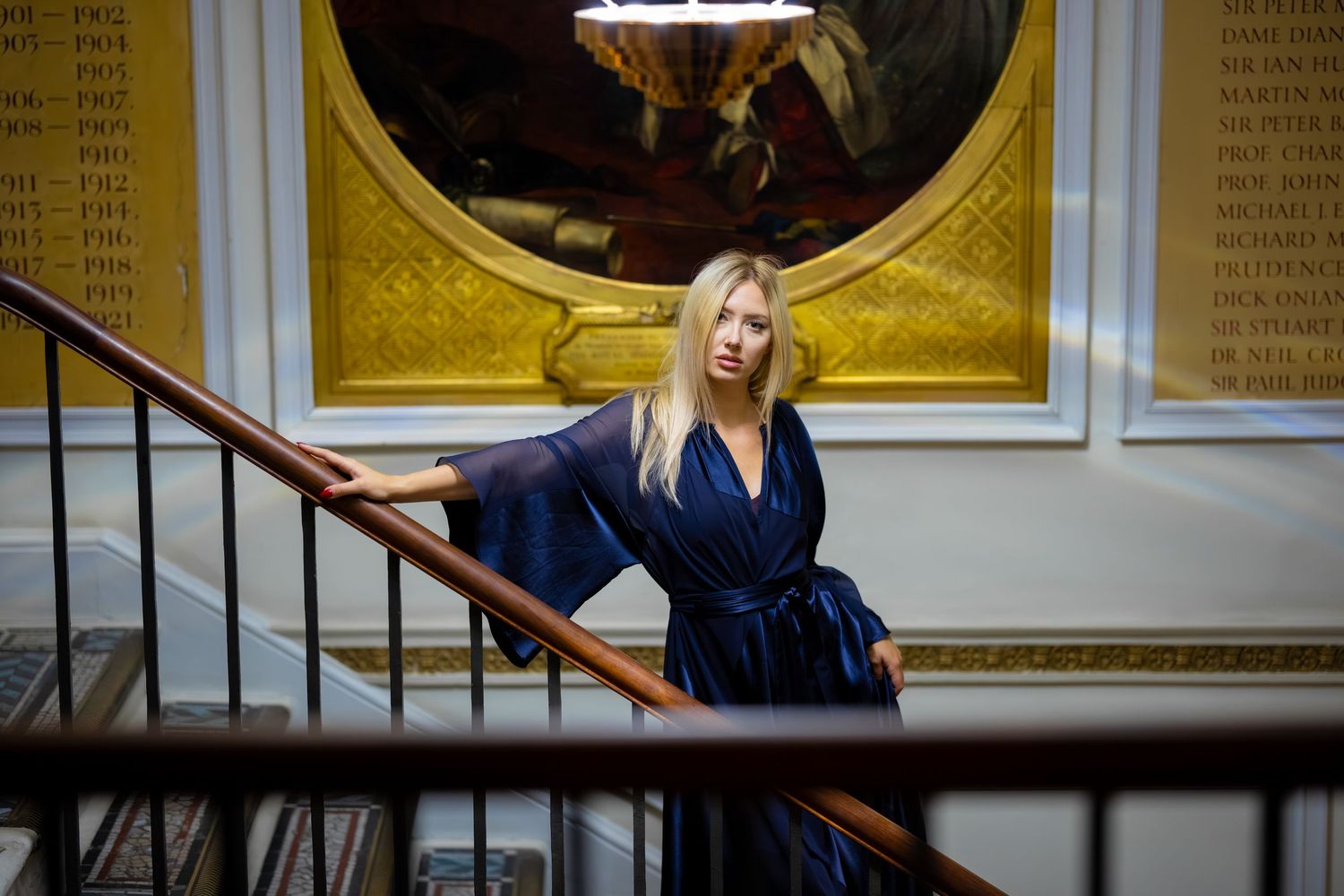 Blonde woman in navy blue wrap dress posing on wooden staircase near ornate gold painting in elegant interior.