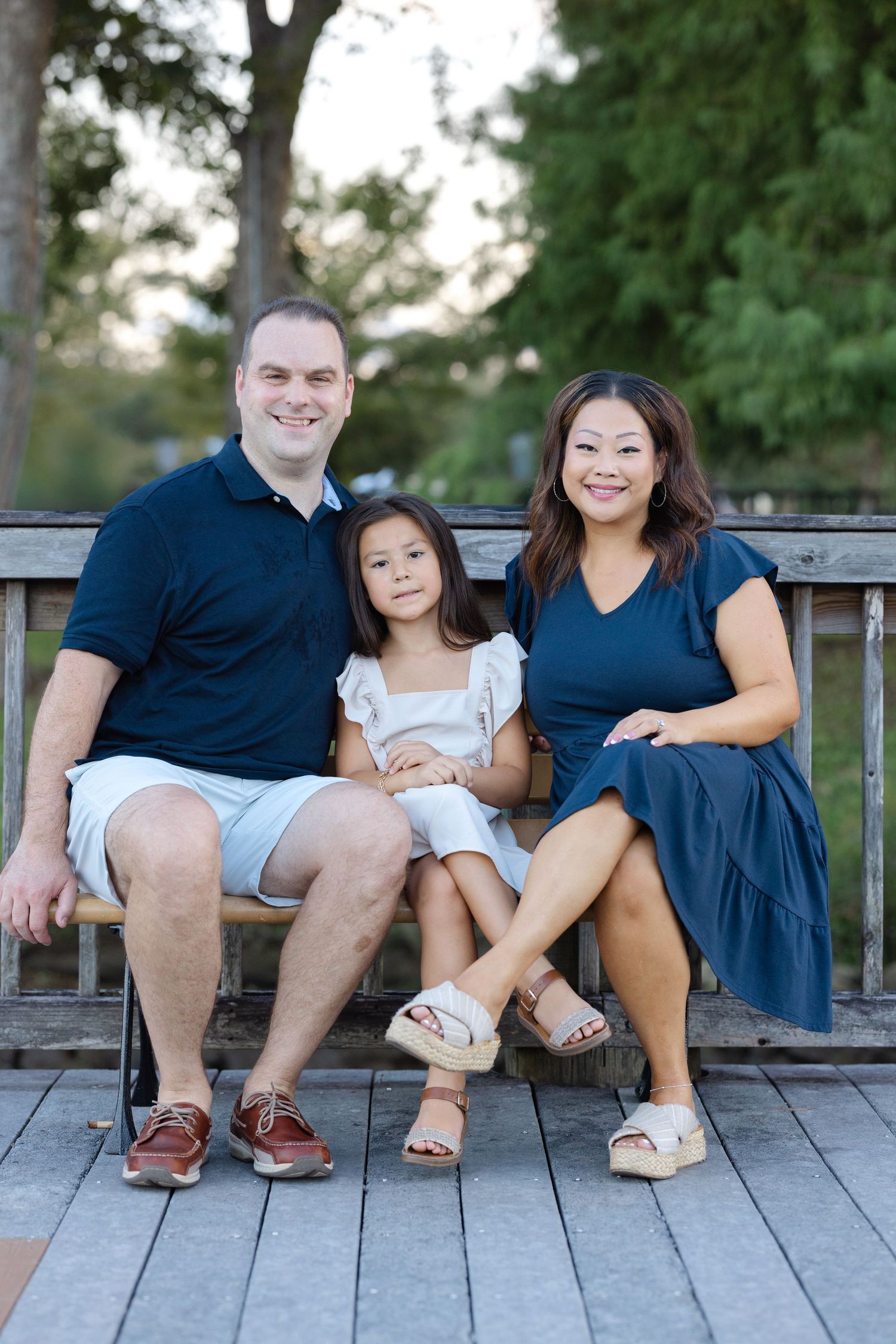 A smiling family of three sits on a wooden bench outdoors, with lush green trees in the background.