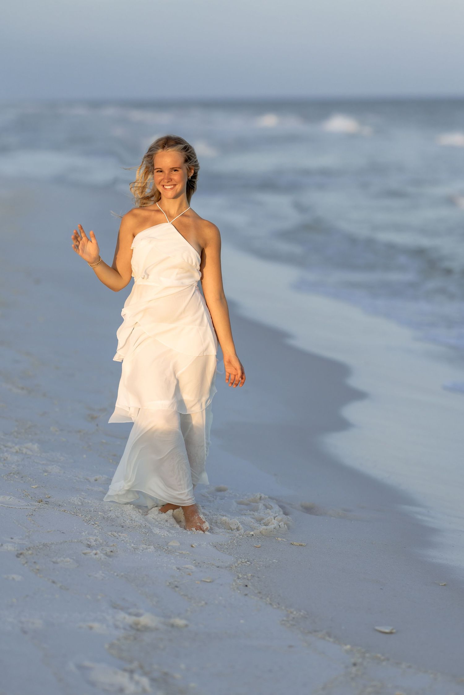 A smiling blonde woman in a flowing white dress walks barefoot along a sandy beach with ocean waves.