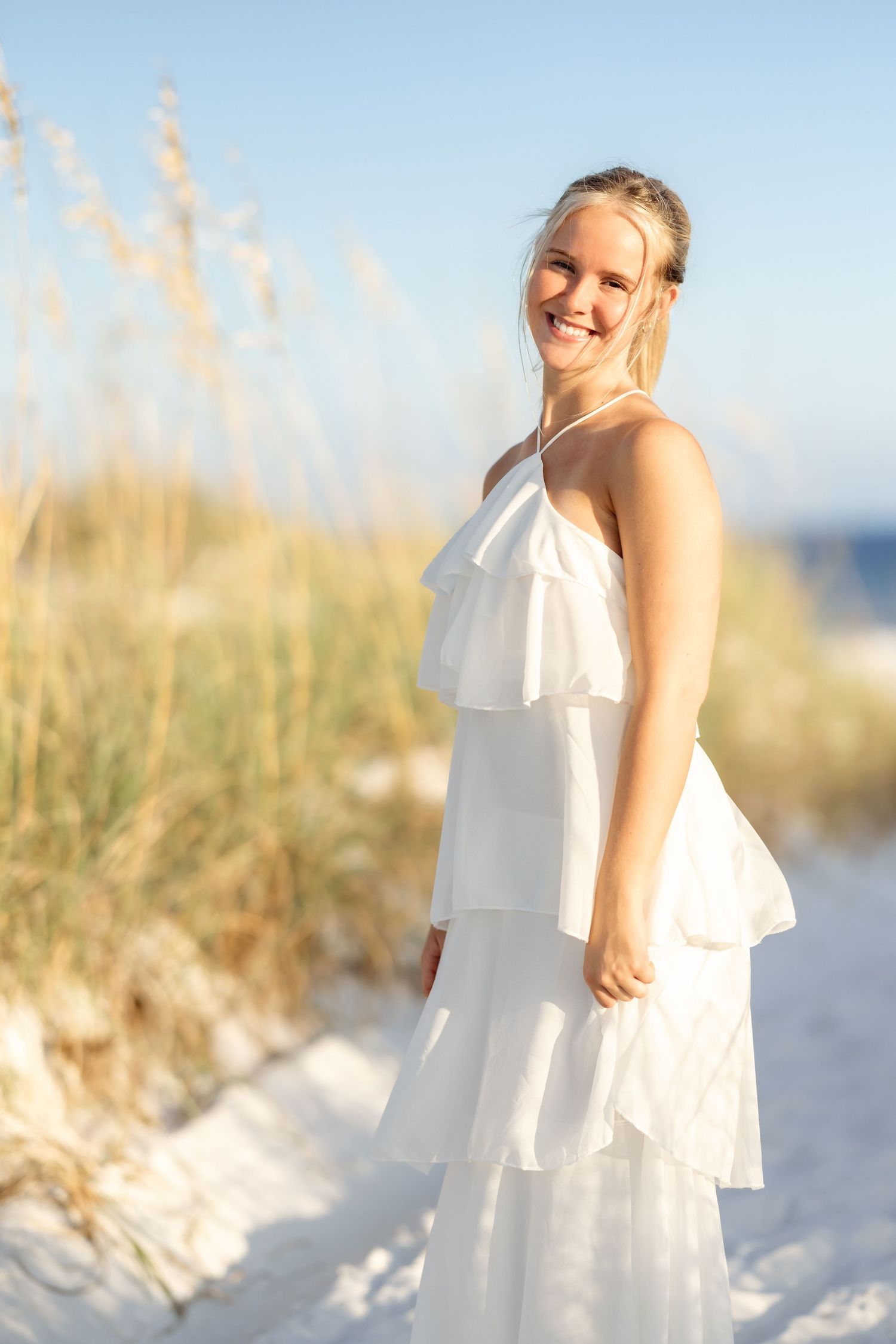 Smiling blonde woman in white ruffled maxi dress standing on sandy beach dunes with sea oats in golden light.