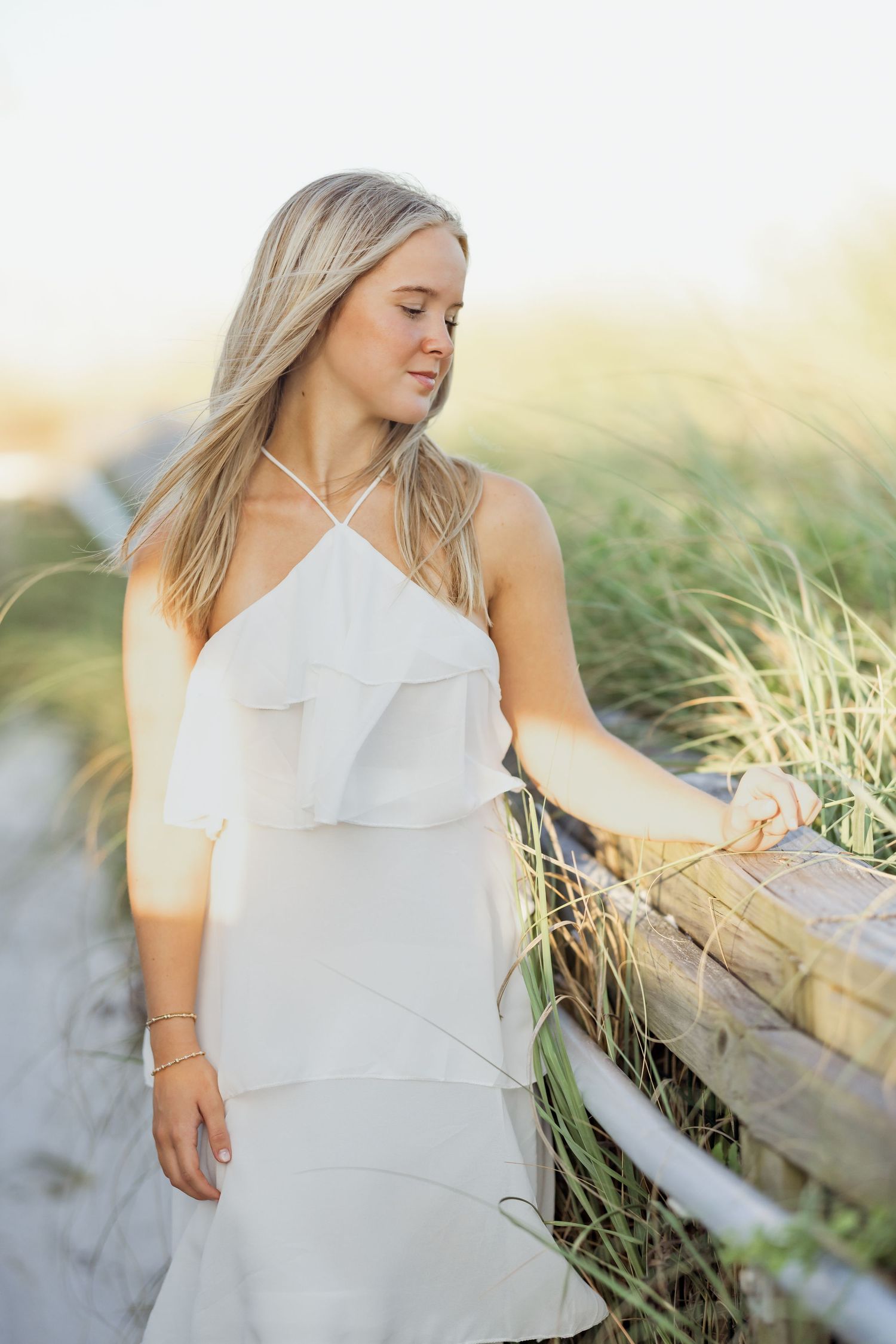 Young blonde woman in white halter dress poses by wooden beach fence with sea grass in golden hour light.