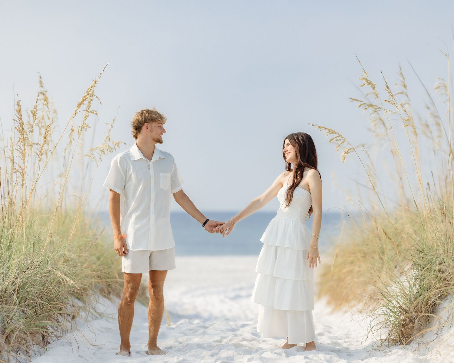 Couple in white outfits holding hands on a sandy beach path surrounded by sea oats.