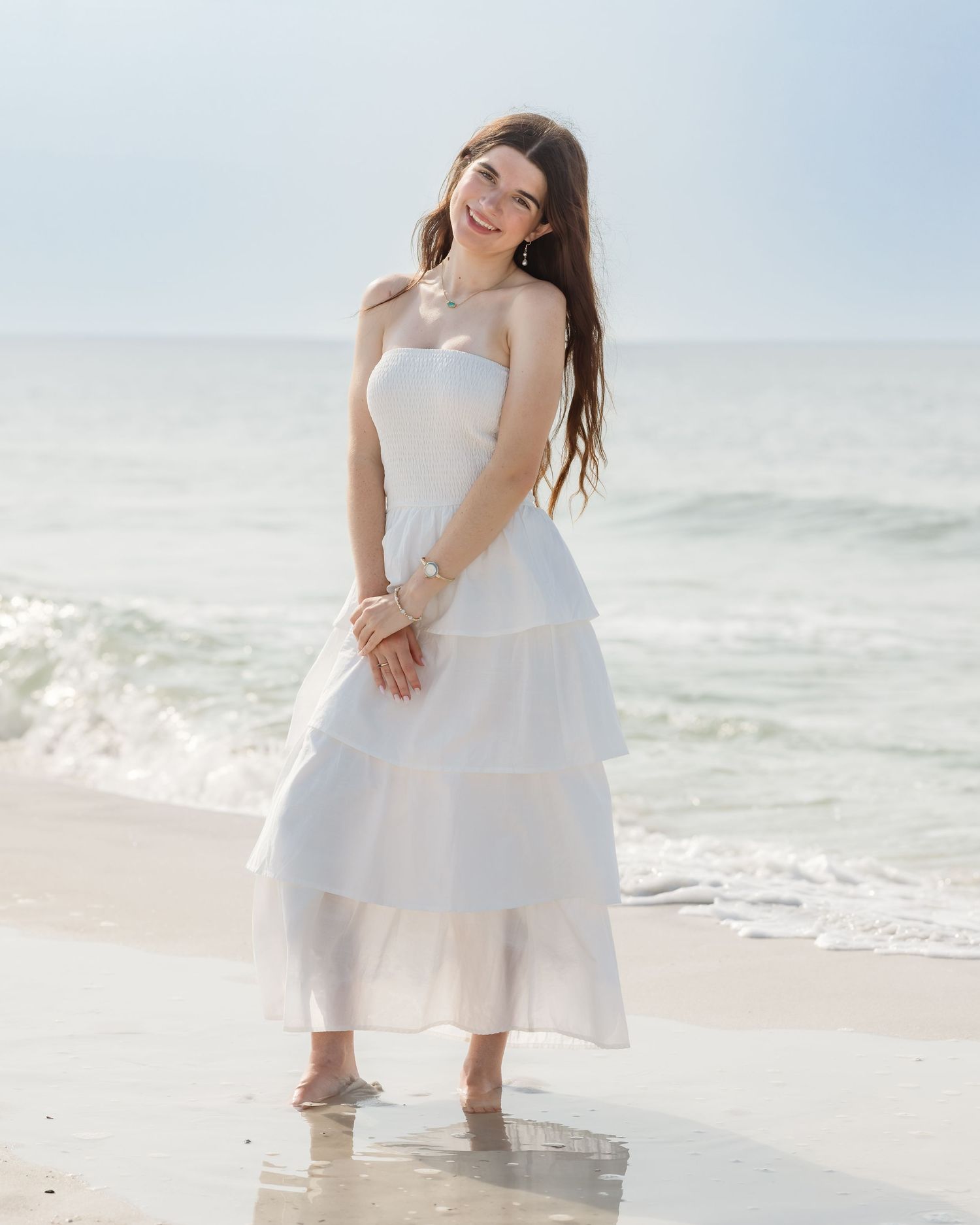 Young woman in white tiered strapless maxi dress smiling on a sandy beach with ocean waves behind her.