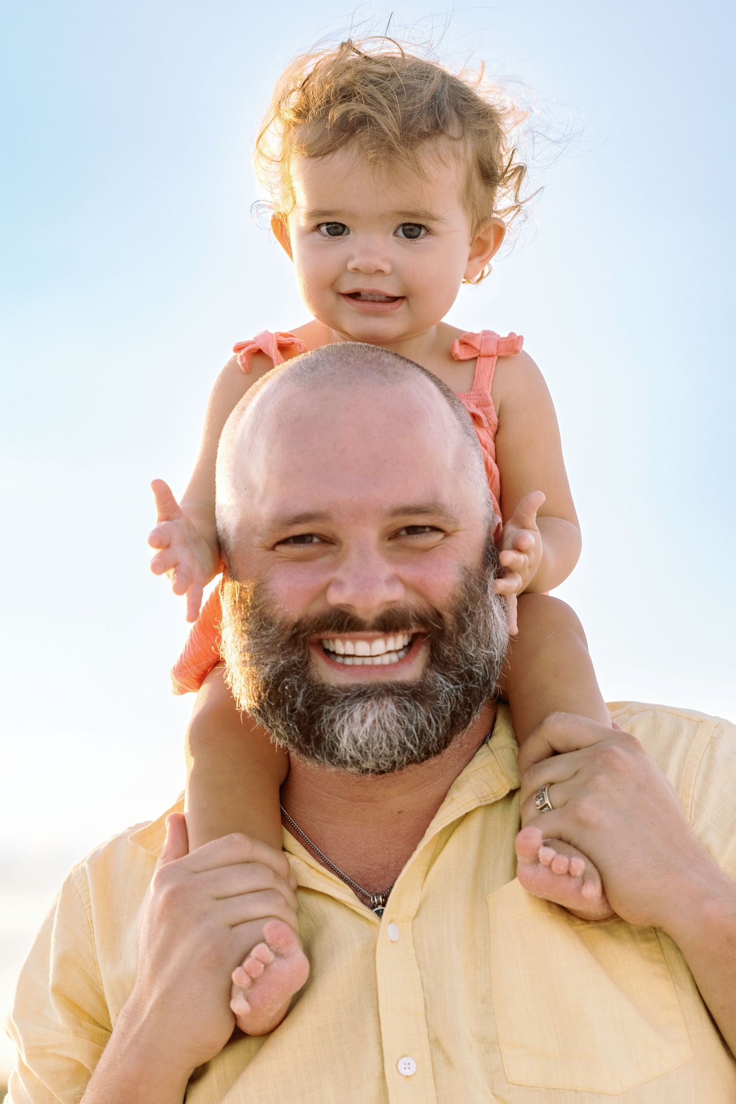 A joyful interaction between an adult with a gray beard wearing yellow and a young child on their shoulders outdoors.