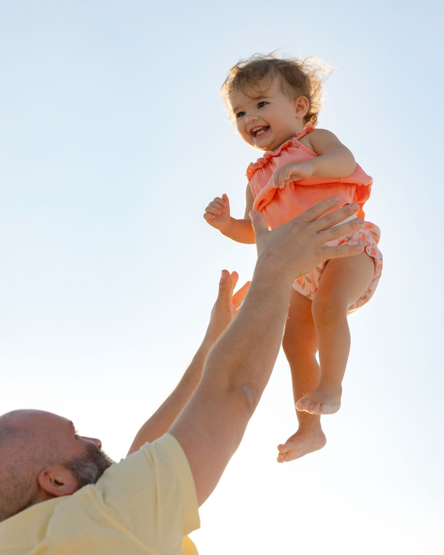 A sequence of photos shows a parent playfully lifting a baby wearing an orange outfit against a bright sky background.