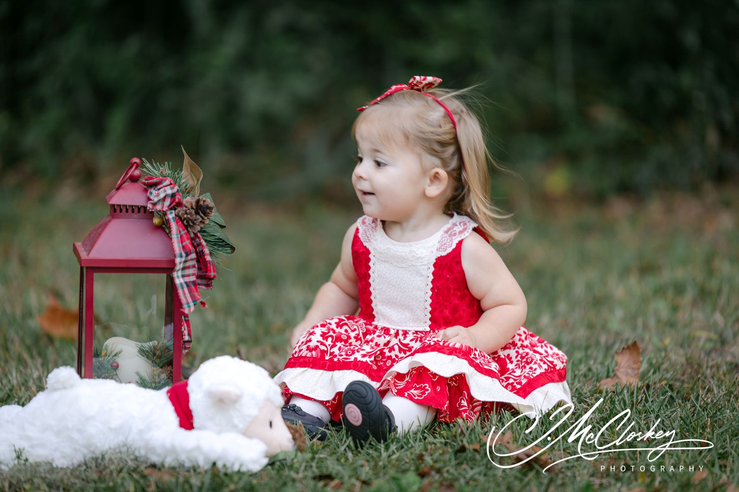 A baby girl in red and white outfit sits and plays in a grassy outdoor setting.