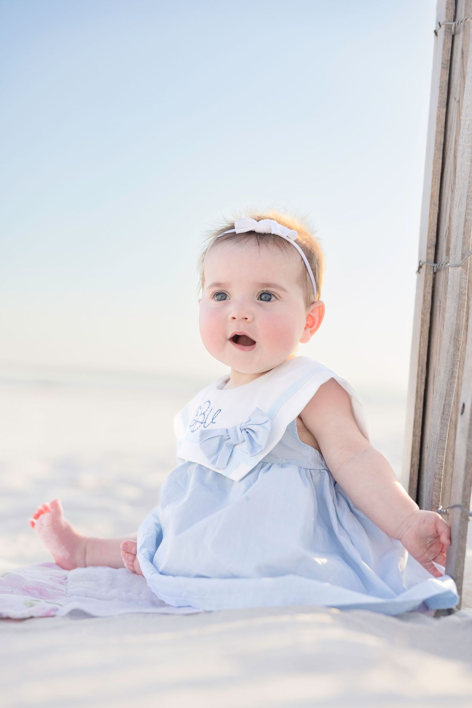 A small child wearing a light blue dress with white ruffles sits on a sandy beach.