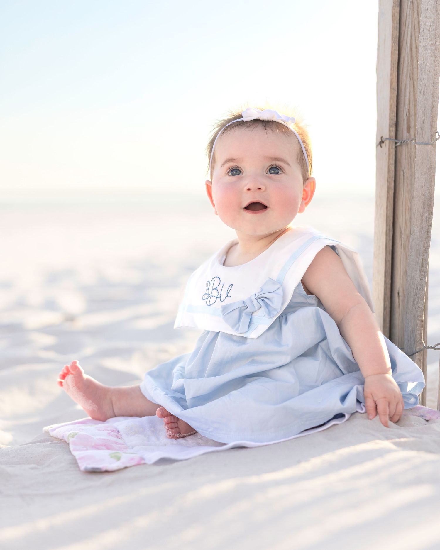 A baby in a white dress sits on soft white sand at the beach during sunset while looking playfully at the camera.