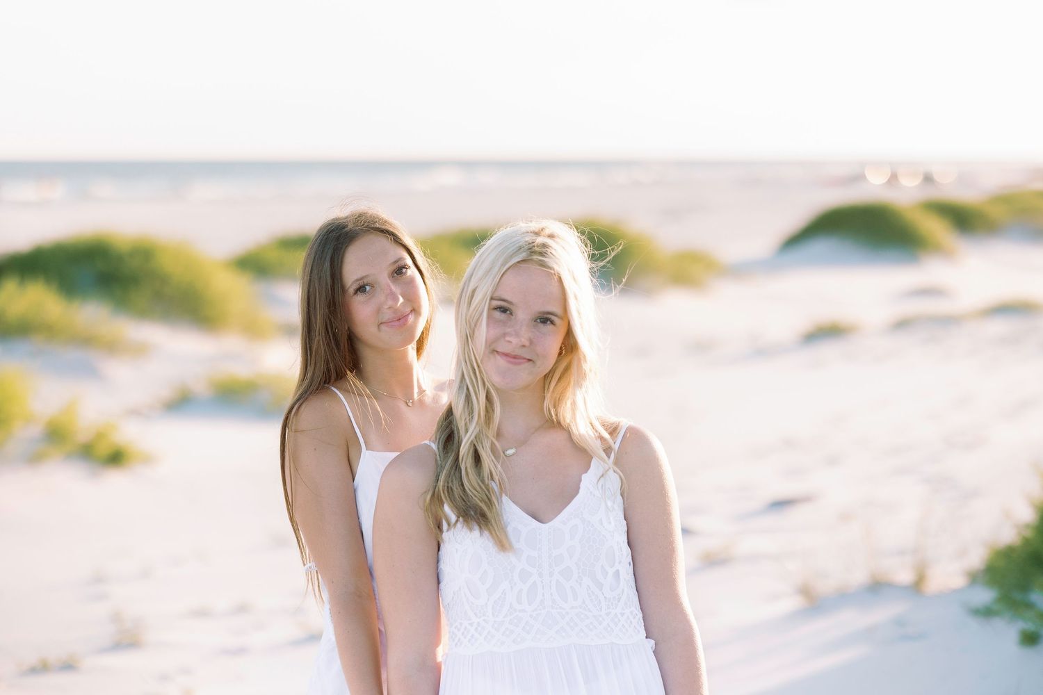 Two women in white outfits walking together on sandy beach during sunset.