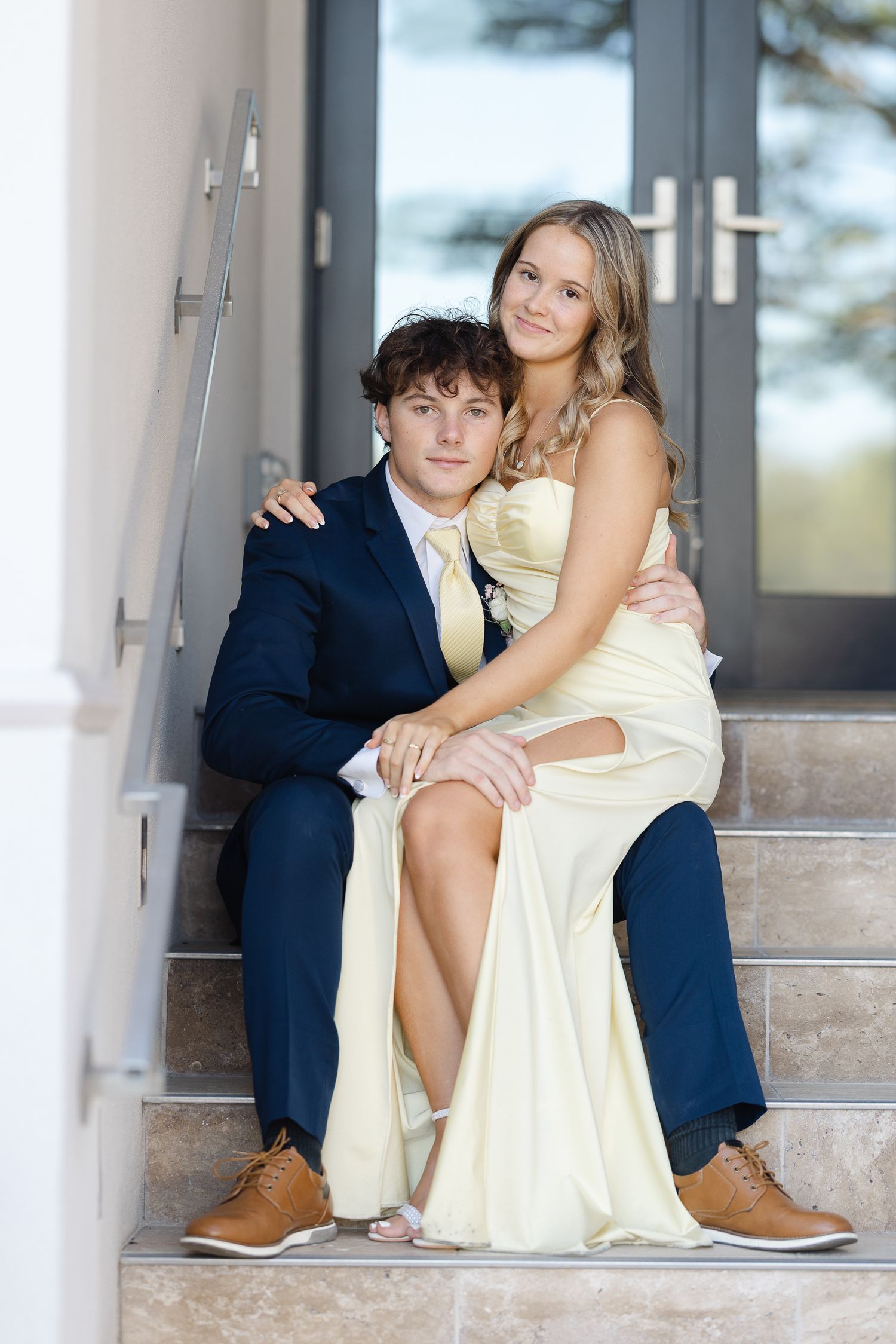 Formal couple in yellow dress and navy suit sitting together on concrete steps.