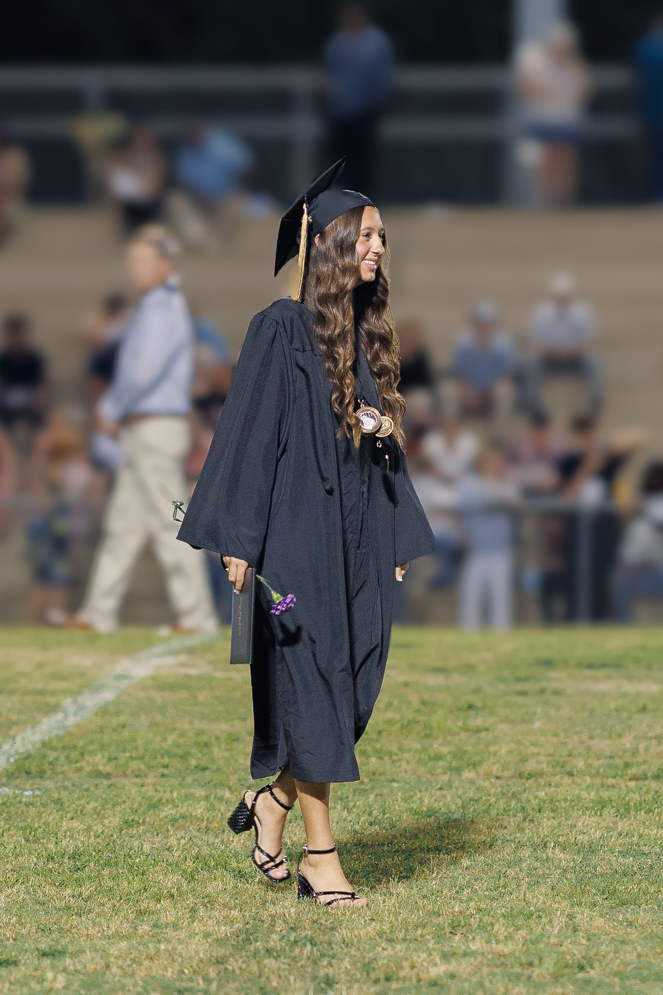 Graduate in flowing black graduation gown walks across field wearing strappy heels.