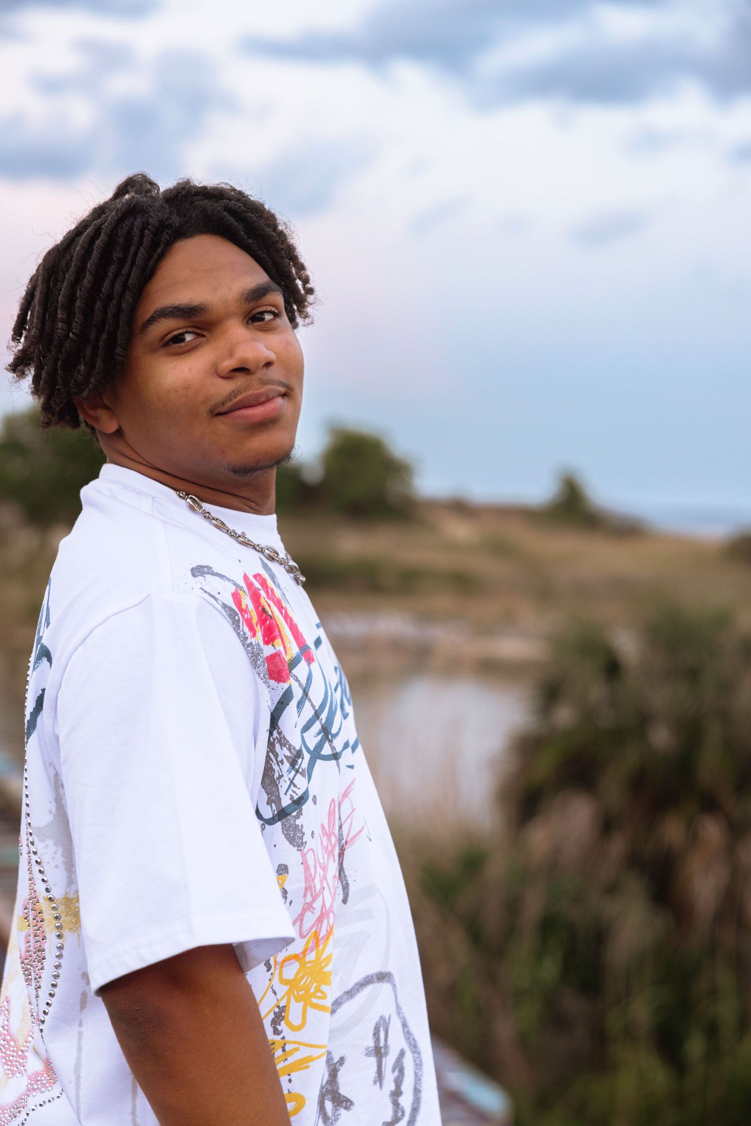 Young man with dreadlocks wearing a white graphic t-shirt poses outdoors with a cloudy sky background.