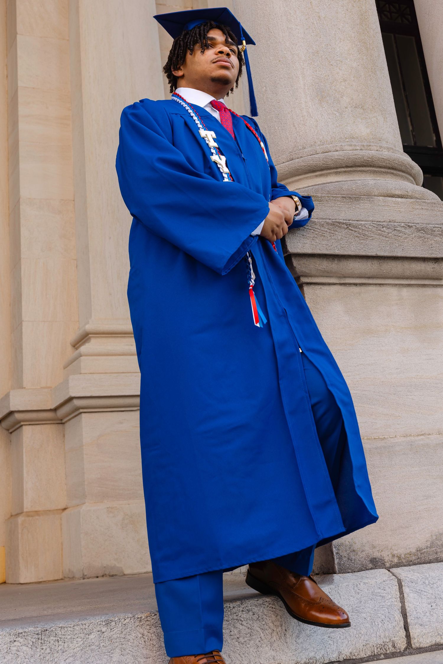 Young man in royal blue graduation gown and cap poses proudly on stone steps of a classical building.