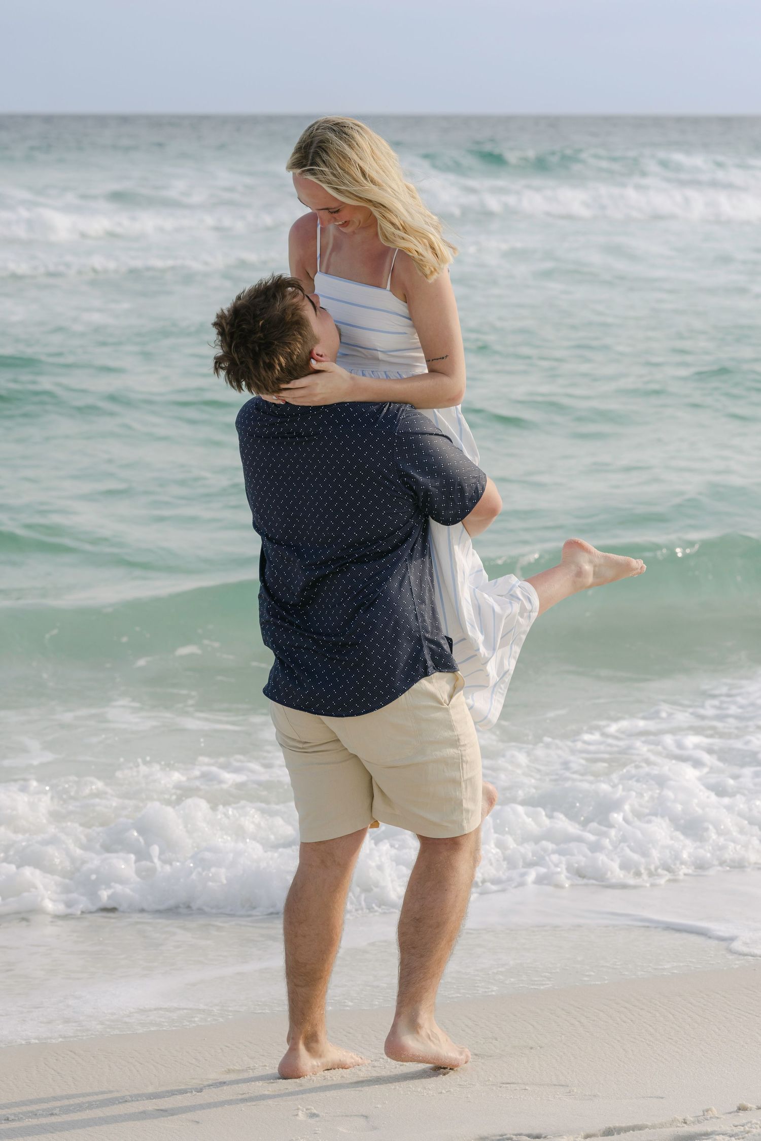 Man lifting woman in his arms on a sandy beach with turquoise ocean waves behind them.