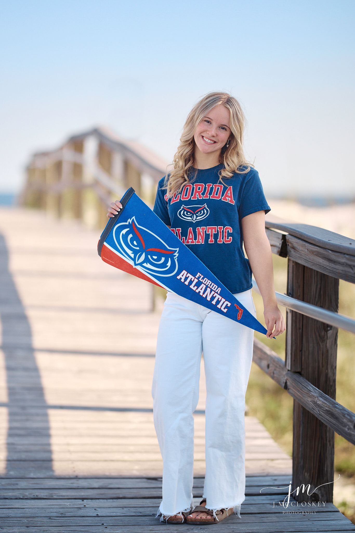 A student in a Florida Atlantic University shirt holds a blue pennant while standing on a wooden boardwalk at the beach.