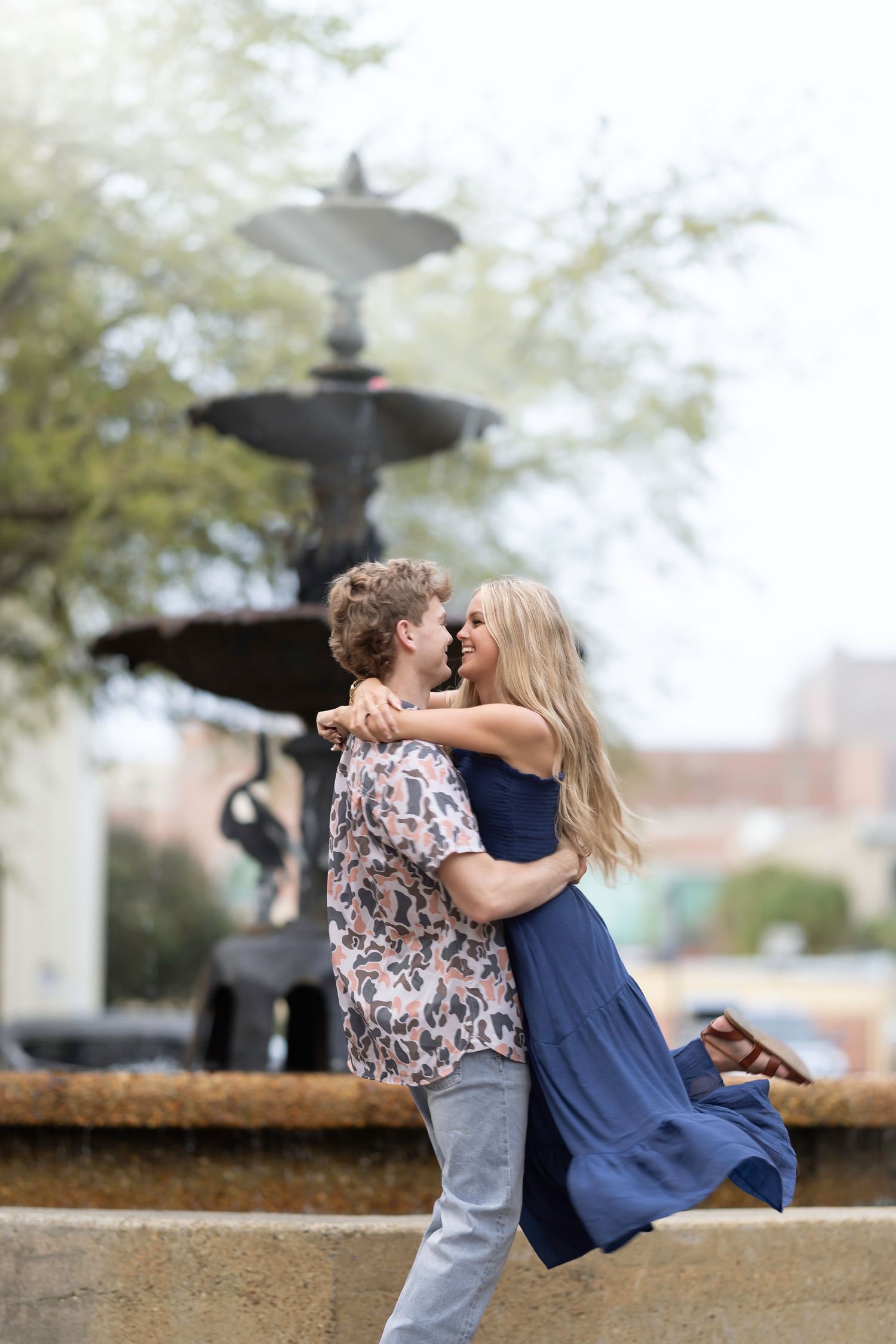 A couple shares a romantic kiss near a fountain in an outdoor setting on a misty day.