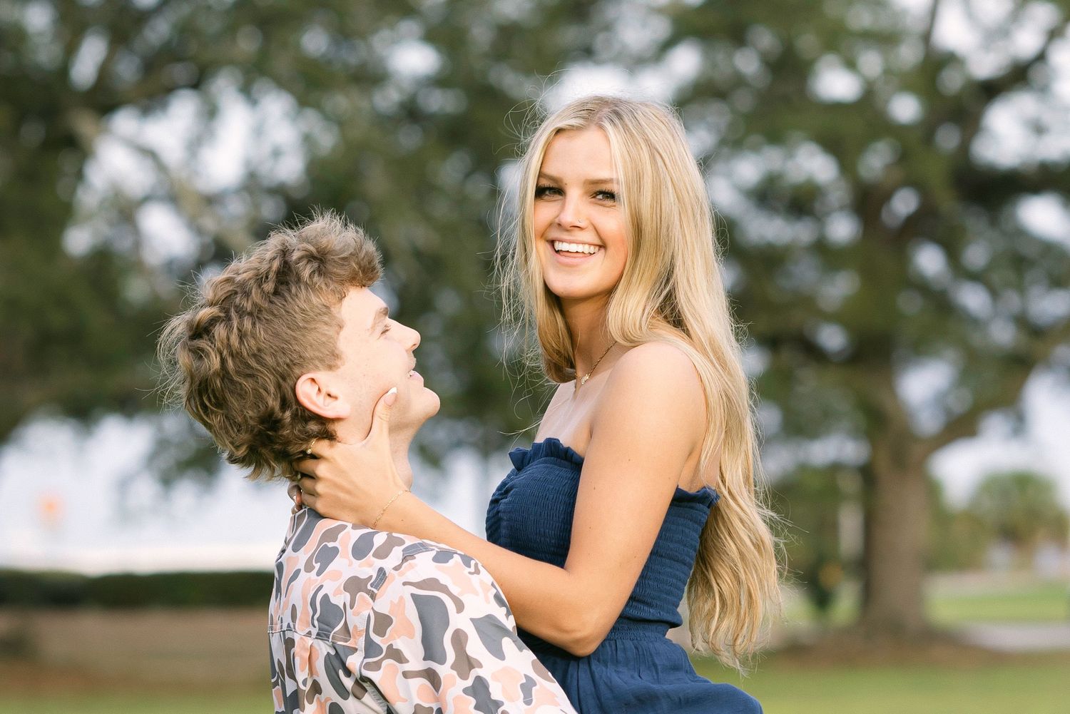 Two people share a joyful moment outdoors in a park setting with green trees in the background.