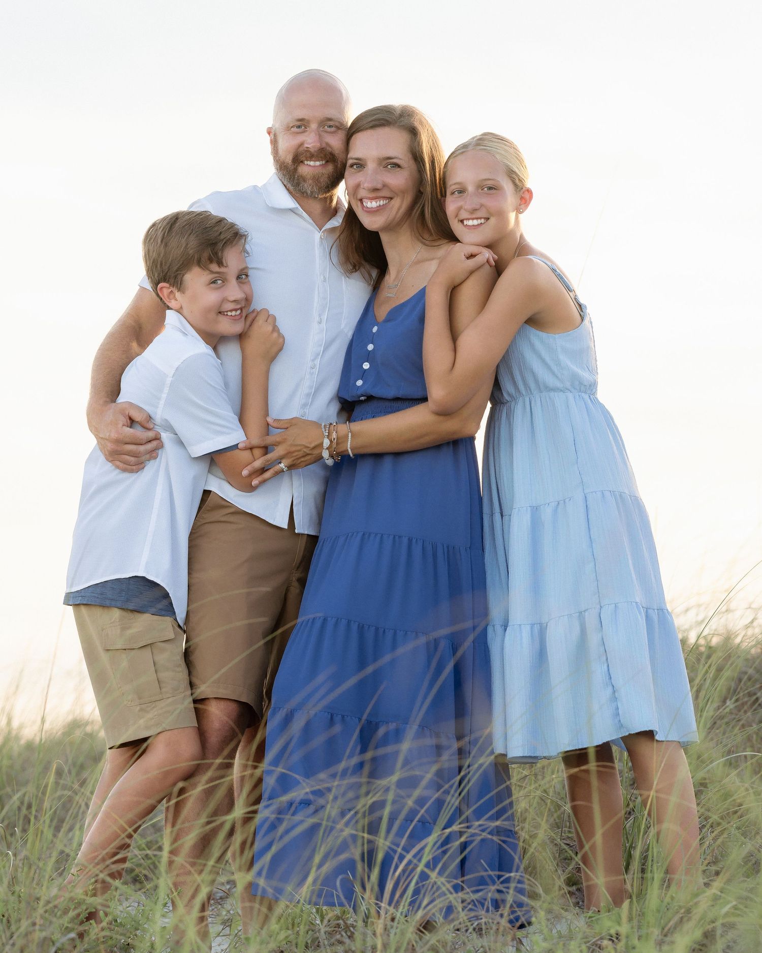 Happy family of four embracing on a sandy beach at sunset, dressed in coordinating blue and white outfits.
